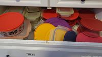 Drawer showing multiple plastic lids and containers of different colors and shapes, stacked and arranged inside a kitchen drawer.