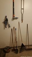 Photo of metal crutches, hand-held weed puller, edger hanging on wall, and several rakes and shovels standing on floor below them, with small basket of hand tools
