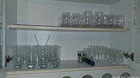 Top shelf and bottom shelf of white cabinet showing various glassware items including tumblers with straws, stemmed wine and champagne glasses, and short rounded glasses.