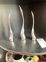Photo of three polished aluminum abstract Hozelton figures with elongated curved shapes on black shelf.