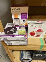 Photo of boxed Veggie Bullet Blender Kit, Angel Hair Blade spiralizer blade boxes, and The Pomegranate Tool box on wood surface.