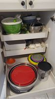 Kitchen cabinet drawer containing stack of stainless steel mixing bowls with red lids, green and white salad spinner, citrus juicer, colorful plastic plate lids, double egg cooker, and clear plastic container.