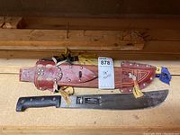 Full view of PROMEDOCA machete alongside its brown leather sheath with metallic rivets and tassels on wooden surface.