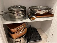 Shelf with stacked stainless steel pots with lids, metal mixing bowls, copper-colored roasting pans, and kitchen utensils including ladles and turners.