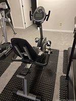 Full side angle of the Schwinn 212 recumbent exercise bike on rubber mats in a basement room with carpet floor.