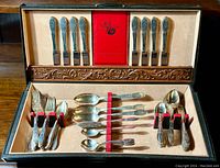 Full view of the flatware set arranged inside the vintage flatware chest with elastic bands securing forks, knives, and spoons. Chest interior lined with beige felt and wood trim with carved design.