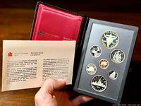 Proof coin set displayed in open black case revealing seven coins and certificate held in hand