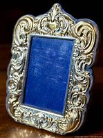Close-up image showing detailed ornate silver-plated repousse design on the photo frame with blue fabric backing visible through glass.