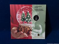 Front of commemorative coin set packaging showing snow globe with Christmas trees and a visible coloured quarter coin.