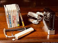 Black Nintendo Wii console on stand with power adapter, sensor bar, composite A/V cable, Wii Remote, Nunchuk and stack of game cases