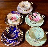 Five Paragon fine bone china teacups and matching saucers arranged in group showing variety of colors and floral patterns including teal, cobalt blue, black, pink, and gilded roses.