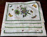 Four white cotton placemats stacked, showing the botanical illustrations and green leaf vine border with butterfly motifs.