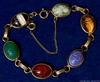 Top-down photo of the bracelet laid flat on dark blue fabric, showing six carved oval scarab gemstones in green, black-and-white banded, light green, amber, purple-blue, and red colors, all set in gold-colored metal links with a spring ring clasp and security chain.