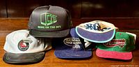 Six vintage caps positioned on wood surface, including black Argos CFL mesh cap with "BRING ON THE 90'S" text, white cap with patch, blue 'National Truck League' cap, cream cap with blue trim, green and red cap, and Starter cap with tags.