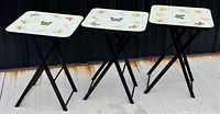 Full view of three vintage folding TV tables with butterfly tops and black lacquered legs displayed outdoors on concrete.