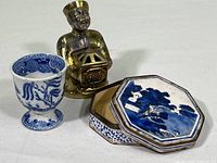 Photo showing the Blue Willow design ceramic egg cup, Chinese brass incense holder shaped as a seated figure, and the 8-sided metal enameled painted covered container with a lid.