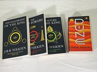 Four paperback books on a flat surface: three volumes of Lord of the Rings in black covers and Frank Herbert's Dune with an orange desert-themed cover.