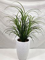 Front view of large artificial plant with many narrow grass-like leaves in tall white textured ceramic pot against white backdrop.