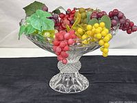 Full view of large clear glass pedestal dish filled with varied colored faux grape bunches.