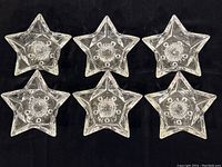 Top-down view of six star-shaped glass candle holders arranged in two rows of three on a black background, showing the clear glass and star design.