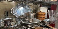 Wide view of multiple metal and wooden barware and serviceware pieces including trays, glass jar with metal lid and handle, wooden jar, and glass tumbler on metal shelf