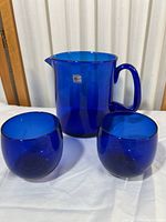 Full set of cobalt blue Blenko glass pitcher with two matching glasses on white surface with wooden background