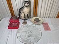 Full lot view including cat pillow, etched glass plate, cat decorative plate, bell, and two boxed glass ashtrays.