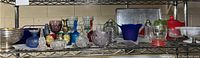 Front shelf view showing various colored and clear glass items arranged on a metal wire shelf including bowls, vases, and decorative glassware