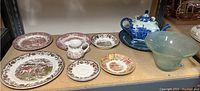Overview of colorful plate collection including a variety of plates, a teapot, and a glass bowl displayed on a shelf.