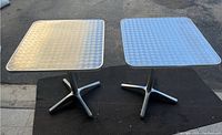 Two commercial stainless steel turn top side tables shown on a black mat on concrete ground, displaying tops and bases with visible surface wear and paint residue.