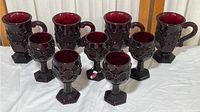 Full view of 9 cranberry colored glassware items, 4 large handled cups and 5 smaller goblets, all embossed with textured circular and geometric designs.