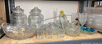 Shelf displaying two large lidded containers, a large cut glass basket, several small glasses, cut glass goblets, a small vase with a flower, a heart-shaped dish, covered basket, and a small blue dish with green glass pieces.