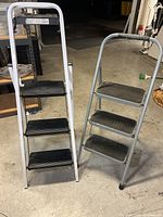 Two folding step ladders side by side showing black textured steps and aluminum frames.