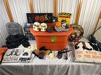 Full display of Halloween decor items arranged on and around a large orange 25 gallon tote including skulls, pumpkins, signs, pillows, gloves and ornaments.