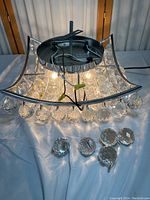 High-end hanging crystal chandelier laid on surface showing curved square metal frame with crystal globe pendants hanging from metal loops. Several crystal globes shown detached below main frame.