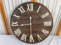 Large round farmhouse style wall clock with wooden face, white distressed Roman numerals, and clock hands showing 3:45. Entire clock face shown.