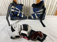Photo showing Nordica ski boots, white and black Hestra ski gloves, and ski goggles all on a white surface.
