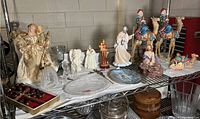 Full view of multiple religious themed porcelain, glass, and metal figurines and decorations arranged on a wire shelf, including large angel, three wise men on camels, nativity scene, and various plaques and figurines.
