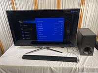 Samsung 55-inch TV on stand, displaying settings menu. Wi-Fi sound bar and subwoofer visible in front and beside TV.