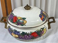 White enamel Dutch oven pot with decorative cornucopia fruit pattern, brass handles, and brass knob on lid.