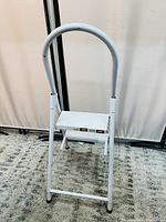 White foldable metal ladder with 2 steps and curved handle, shown unfolded on carpet.
