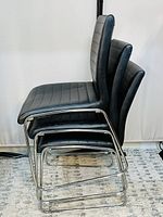 Side view of three stacked black leather finish chairs with chrome metal frames on carpeted floor against white backdrop.