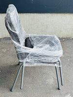 Side view of one black cushioned chair wrapped in clear plastic showing metal legs and padded armrests.