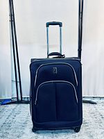 Front view of black fabric suitcase with two front zippered pockets, telescopic metal handle extended, four wheels on bottom, standing upright on carpeted floor.