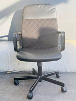 Front view of black office chair showing padded seat, diamond quilted backrest, armrests, and caster wheels on base.