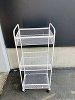 Front view of white metal rolling storage cart showing three mesh baskets on caster wheels against a black and gray wall background.
