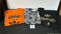 Three folded Adidas shirts side by side - orange with logo, camouflage, black with gold print