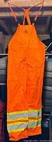 Full view of the bright orange bib pants showing reflective bands on the leg cuffs and adjustable orange shoulder straps.