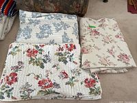 Two floral pattern pillowcases and a medium-sized vintage floral blanket on carpeted floor