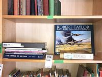 Shelf with several aviation books including 'The Battle of Britain' by Jon Lake, 'Air Combat Paintings Volume III' by Robert Taylor, and other WWII aviation titles.